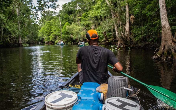 From the back of a canoe, you can see the people in front paddling. The boat is on calm water with trees lining the shore. 